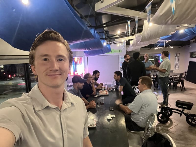 Rand Larsen taking a selfie at a networking meetup in Atlanta, with a group of attendees seated and standing around a long table in a decorated venue