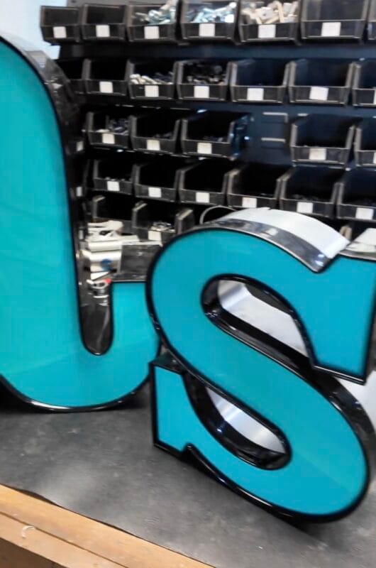 Large teal and black dimensional letters spelling US on a workbench, with a wall of parts bins in the background