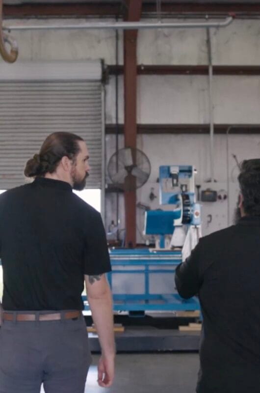 Two men in black polo shirts viewed from behind, looking at a large blue milling machine inside a CLN of South Florida manufacturing facility